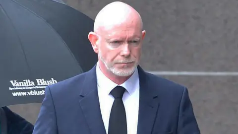 Philip Braat, a bald man with a well-trimmed grey beard and moustache, walking towards a court building. He has a serious expression and is looking at the ground. Braat is wearing a navy suit, white shirt and a black tie