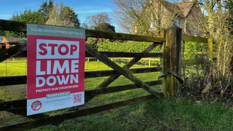 A wooden field gate with a 'Stop Lime Down' poster in red and white block capitals attached to it. Through the gate can be seen green fields behind.