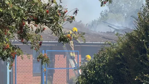 Jamie Niblock/BBC Firefighters attending a blaze at Thomas Bullock Primary Academy. The picture has been taken from a vantage point between an overhanging tree and a leafy green bush. Two firefighters are on a ladder at the side of the school building. There is damage to the roof, which is charred and exposed. Smoke is coming off the roof.