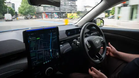 PA Media The hands of a driver can be seen hovering near a steering wheel as a car is in motion, indicating that a human driver is present but not in control.