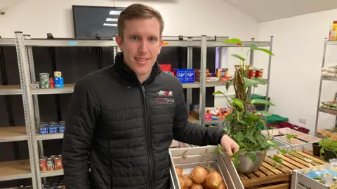 John Gilbert looking into the camera while holding a small crate of onions. He is standing in front of shelving on which there are cans of food.