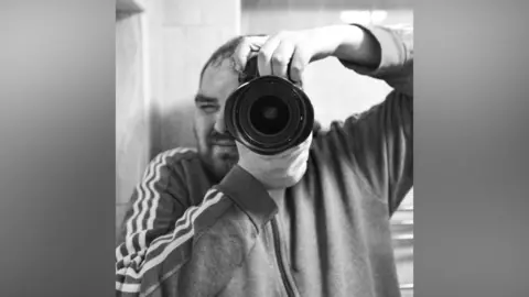 Neil J Catley A black and white image of Neil Catley. He has and beard and short dark hair and is holding a camera up to a mirror to take a photograph. He is wearing a grey zipped top with stripes down the arms. 