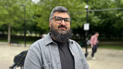 A man wearing glasses and a grey shirt over a black top standing outdoors near a park, with trees, a paved walkway, and a passer-by in the background