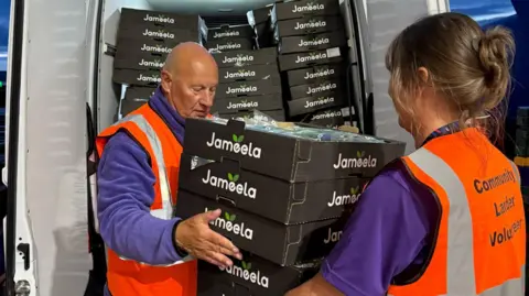 Towcester Community Larder A woman passes a pallet of food to a man who is loading it into a van. They are both wearing hi-vis jackets.