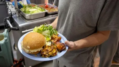 A prisoner dressed in grey is holding a plate with a salad, burger buns and pieces of chicken. He is stood in a kitchen, with food in stainless steel containers on a hot tray behind him. 