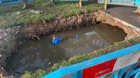 Colin Pattison A hole in a pavement filled with water and with a pipe exposed. It is surrounded by grass and blue barriers.