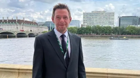 David Williams, wearing a suit, stands by a river. Buildings can be seen on the other side of the water.