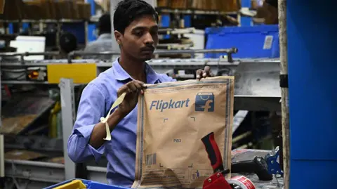 Getty Images An employee in a light purple shirt seen holding paper package with a customer's order. The package has Flipkart's blue logo on it.