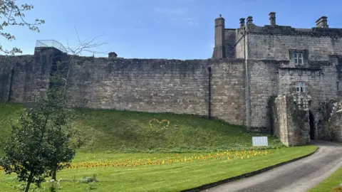Hospice at Home West Cumbria A partial view of the entrance to Cockermouth Castle, with high sandstone walls to one side of a high gatehouse. On the grassed area to the side of the entrance drive are planted dozens of metalwork sunflowers, with more displayed in the shape of a heart on the grassy bank below the walls.