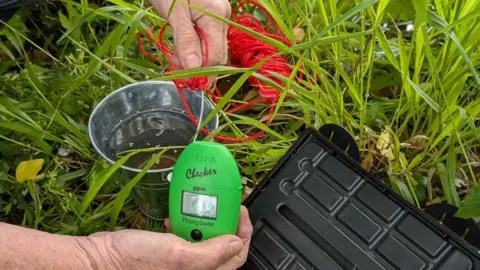 Ruth McDonagh The image shows some of the kit being used to measure different substances in the water. Hands of a man are visible holding a green, handheld, Hanna branded phosphate measuring device. A small silver stainless steel bucket with red string attached to it and a black plastic case for the kit are in the background as well as grass and foliage on the ground.