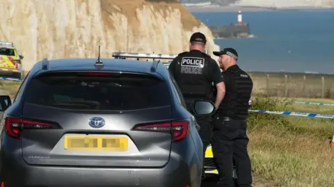 Sussex News and Pictures Two police officers stood next to a car in a field. The field overlooks a cliff edge, with sea in the background and a lighthouse on a jetty.