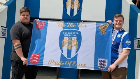 Contributed Mitchel Wraight holds a Colchester United flag with another man holding the other side. Mitchel has short dark hair and wears the club's shirt which is blue and white.