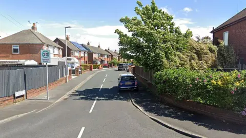 The start of Stainforth Avenue, Bispham, Blackpool. A mini is parked on the right of the street in the shadow of trees from a garden. It is a sunny day. There is a 20 mph road sign saying 'Twenty's Plenty'.