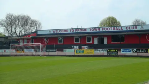 A football pitch with the club building behind it. The building is painted red and says Welcome to Frome Town Football Club on the roof.