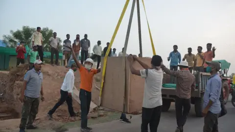 Bhakarba Thakor A crane was used to transport the wooden box containing the skeleton to its present site. The image shows men engaged in transporting the skeleton, surrounded by onlookers.