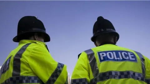 Two police officers wearing a bright yellow jacket with the words POLICE  in bold white lettering in a blue rectangle 