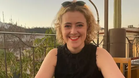 Meg Houghton-Gilmore Meg has short blonde hair and has sunglasses sat on top of her heard while smiling at the camera. She is sat at a restaurant table outside.