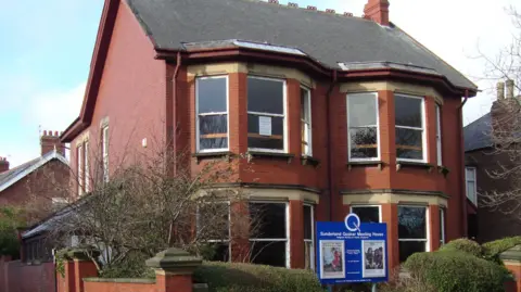 Sunderland Quakers A large two-storey Edwardian house has a blue sign in front of it which says Sunderland Quaker Meeting House. The building has bay windows on both the ground floor and the first floor with a lean-to extension on one side and bushes in the front.