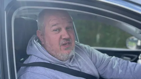 A man wearing a grey hoodie sits in his car with a seat belt on