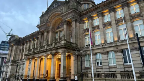 The front of Birmingham City Council house, taken from a slight angle. Four flags can be seen on tall poles. The sandstone building has three floors, with a higher rotunda level, columns holding up a balcony and a central arch.