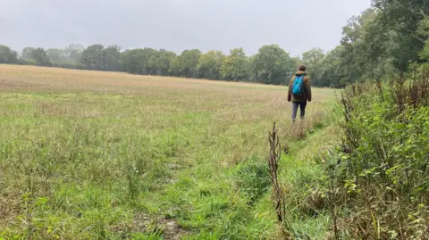 A man walking through a field. He is wearing trousers, a brown coat, and a blue backpack. The field is large and green with foliage and trees to the left and in the distance. The sky is grey.