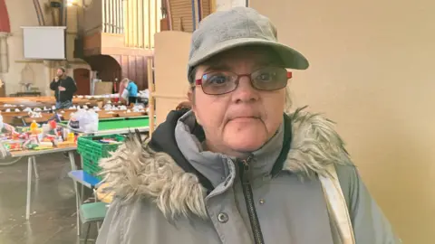 A woman wearing a grey winter coat, cap and red glasses looks into the camera. Food parcels can be seen in the background.