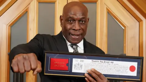 PA Media A man in a suit is holding a formal certificate in one hand, while his other hand is bunched in a fist pointing towards the camera