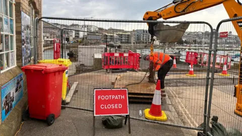 Vanessa Thomas A digger with a chain hanging from it is handled by a person in a high-viz orange jacket while a metal gate in front of him is bordered by a red and white sign which reads Footpath Closed. 