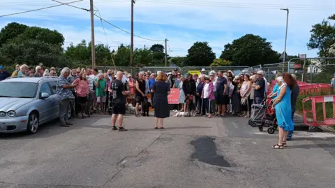 A gathering of people at the site of a mineshift collapse in Troon, Cornwall. They are protesting and calling on Cornwall Council to fill the hole. Metal fencing and a road closure sign are at the site. A silver Jaguar car is parked on the left-hand side.
