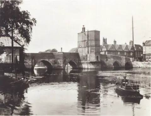 Norris Museum, St Ives Black and white photo of the bridge and chapel, with two extra storeys on the chapel when it was used a residential home, a man in a flat cap and suit can be seen rowing a boat on the right and buildings can be seen behind the structure that stretches across a river.