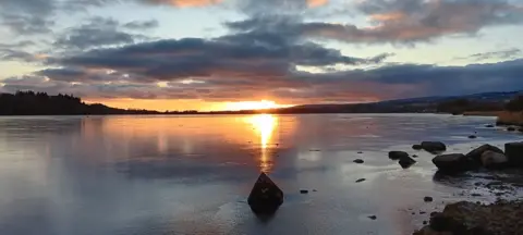 Millar Leon Sun setting on a frozen loch with boulders peeking out of the ice