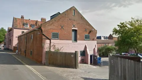 Front of a long building that is made of red bricks and painted pink at the front. There are two fences leading to a car park with trees. 