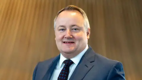 Darren Millar wearing a blue suit, a white shirt and tie. He is photographed in front of the Senedd's central funnel. 