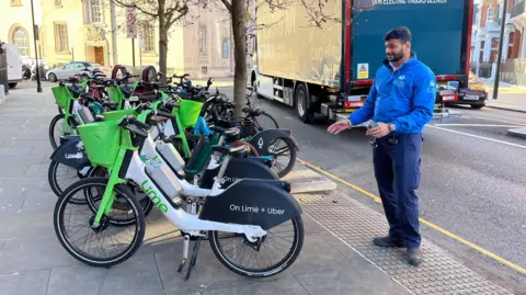 A man in a blue anorak is pointing at a Lime e-bike which is parked in front of a ramp. It is blocking the ramp. 