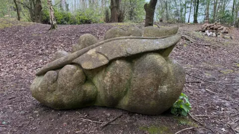 A sculpture made of stone, which appears to be a feather floating on a cloud. It is situated in a woodland with trees and a pile of logs visible behind.