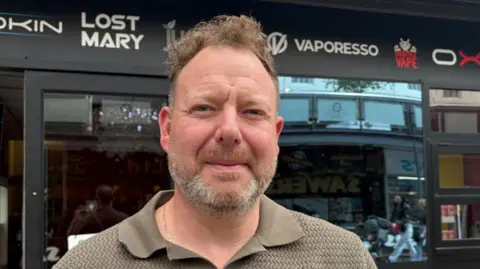 Mr Lagan has short brown hair and a grey and brown beard and moustache. he is wearing a brown polo shirt. He is standing outside his vape store which is black with the brands on it.