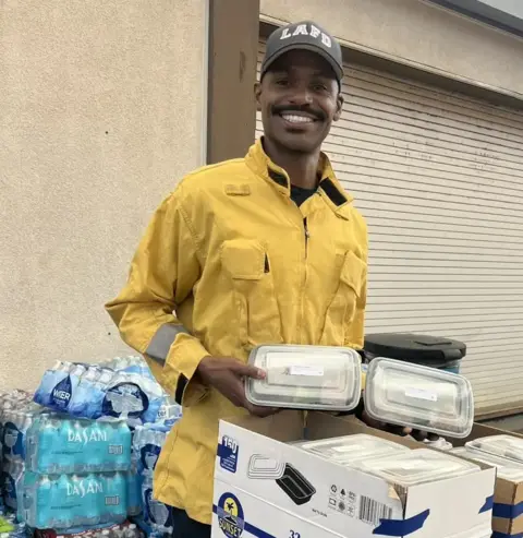 Fardad Khayami A man in a yellow jacket holds several trays of meals, while wearing a Los Angeles Fire Department hat, standing in front of a box of bottled water