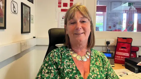 Carolynn Southcombe smiling at the camera in her office. She has brown shoulder-length hair and is wearing a green flowery top.