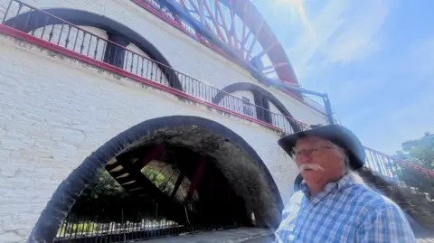 Brian wearing a cowboy has, sits in a wheelchair infront of the Laxey wheel, he was a white moustache and chequered shirt on