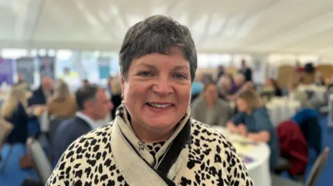 Louise Skelly, a woman with short, dark hair, smiles at the camera in a white marquee tent. She is wearing a cream woolen-type top with a black animal print pattern and a matching cream and black scarf. People are sitting a round tables in the background.
