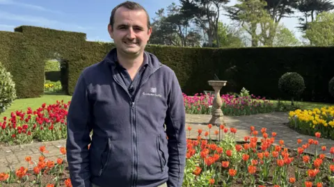 Jordan McWhirter has short brown hair, a navy fleece and navy polo shirt. He stands in the castle gardens in front of a display of tulips. 