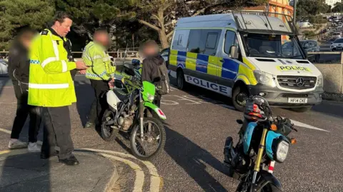 Dorset Police Police officers in high-vis coats and two motorbikes stopped in the street. Two people are standing by with their faces blurred. A police can is parked across the road.
