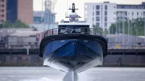 A city skyline with buildings and white sky in the background. The focus of the photo is on a blue boat that appears to be skimming above a river, towards the camera. 