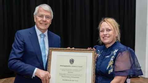 Huntingdonshire District Council Sir John Major wears a navy suit and light blue tie. He is holding one side of a large frame with a certificate inside. The other side is held by a woman wearing a navy dress with a floral pattern on the shoulders.