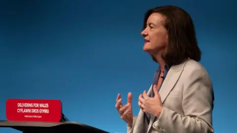 Eluned Morgan stood at a podium delivering a speech at a party conference. She is wearing a beige blazer and is gesturing with her hands. 