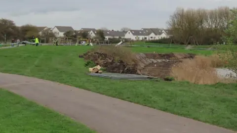 Burnham-On-Sea.com A section of a park is dug up and there are metal barriers and cones on the grass. In the distance is a police cordon and police officers.