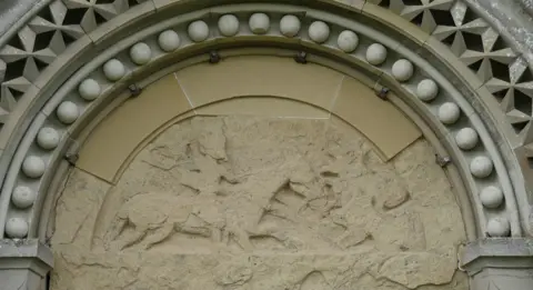 James Towill A semi-circular stone carving feature a man on horseback attack a serpent-like creature