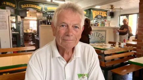 A man in a white polo shirt and white hair short and up on end smiling in a cafe.