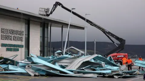 A crane carries out repair work on Helensburgh Leisure Centre - a large building with parts of the roof lying next to it, having been damaged by a storm.
