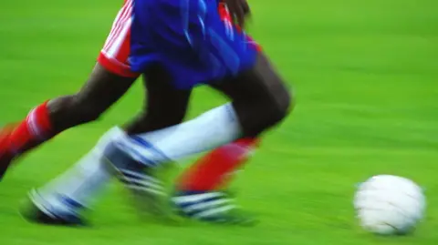 A blurred photo of two football players, playing football, moving forward to kick a white ball